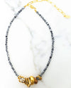 A necklace with black and white patterned beads and a gold chain extender, featuring a cluster of gold rings, a bronze bead, and a silver charm at the center, displayed on a white marble surface.