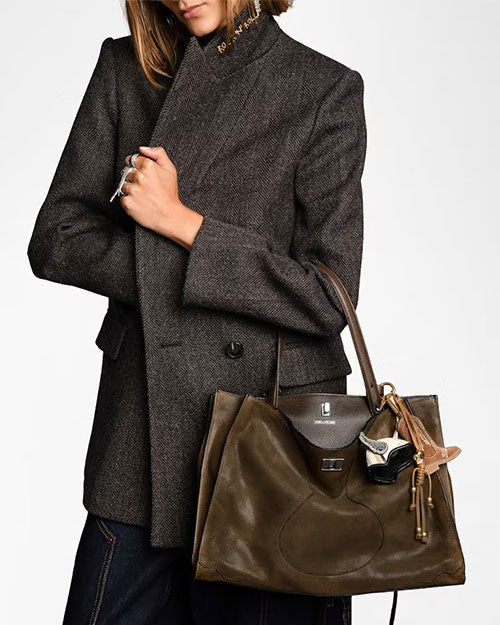 Person wearing a tailored dark gray herringbone blazer, holding a large olive-green leather tote bag with structured handles. The bag features a decorative keychain with a small mask charm and tassel accents.