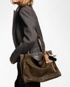 Side view of a person wearing a dark gray herringbone blazer, carrying a large brown leather tote bag with structured handles. The bag features a decorative keychain with a small mask charm and tassel accents.