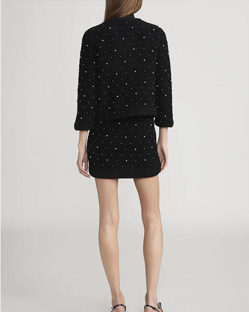 Back view of a black textured two-piece outfit featuring a cropped jacket and a mini skirt. Both pieces are embellished with small pearl-like beads arranged in a scattered pattern. The jacket has three-quarter sleeves and an open front design.