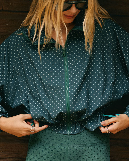 Close-up of a person wearing a teal zip-front jacket with a white polka-dot pattern, matching patterned leggings, and dark sunglasses, adjusting the hem of the jacket against a dark wooden background.