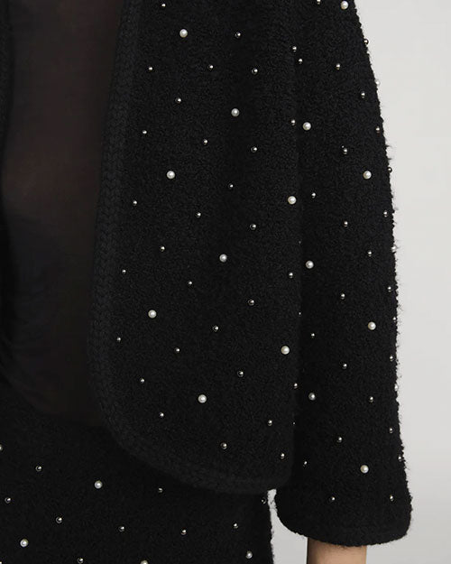 Close-up of a black textured jacket sleeve and part of the front, embellished with small pearl-like beads arranged in a scattered pattern. The fabric has a boucle-like texture, and the edge of the open front is visible.