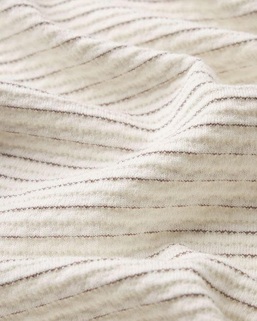 Close-up image of cream-colored fabric with a soft, ribbed texture, featuring fine horizontal lines in subtle tonal variation. The material is gently folded to highlight the drape, depth, and plush surface of the textile.