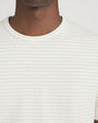Close-up studio image against a light neutral background, showing a cream short-sleeve crewneck T-shirt with fine horizontal ribbed texture. The image focuses on the neckline, upper chest, and shoulder area, highlighting the subtle tonal stitching and fabric detail.