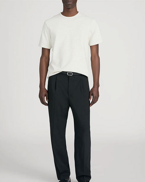 Person standing in a studio against a light neutral background, wearing a cream short-sleeve crewneck T-shirt with subtle horizontal ribbed texture, paired with dark tailored trousers and a black belt. The outfit is styled with black shoes, and the image is photographed in a full-length, front-facing pose.