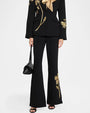 Black tailored suit with gold floral embroidery on the blazer and flared pants, paired with a gold neck scarf, black handbag, and open-toe heels.