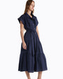 Person standing in a studio against a white background, wearing a navy blue midi dress with short sleeves, a button-front design, a cinched waist tie, and a tiered skirt with subtle textured detailing. 