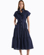 Person standing in a studio against a white background, wearing a navy blue midi dress with short sleeves, a button-front design, a cinched waist tie, and a tiered skirt with subtle textured detailing. 