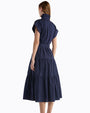 Back view of  a Person standing in a studio against a white background, wearing a navy blue midi dress with short sleeves, a button-front design, a cinched waist tie, and a tiered skirt with subtle textured detailing. 