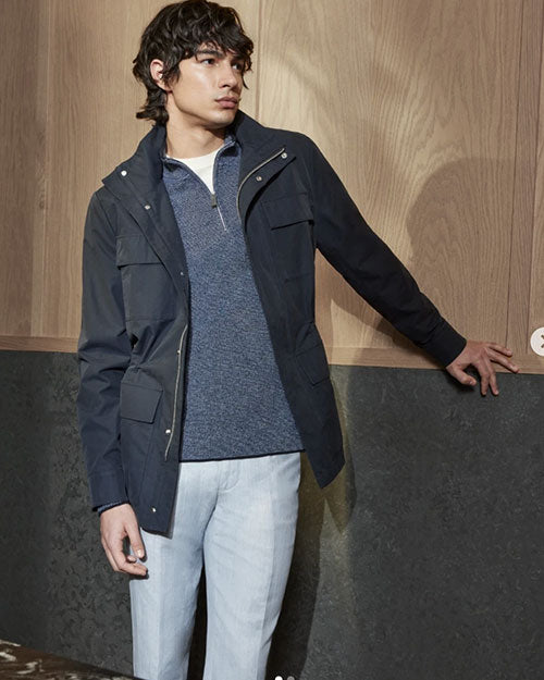 A person stands indoors against a wall with wood paneling and dark textured material, wearing a navy utility jacket over a blue zip-up sweater and light-colored pants.