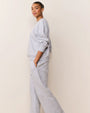 Side view of a person wearing a light gray matching loungewear set with a subtle embossed pattern, featuring a long-sleeve sweatshirt and relaxed-fit pants with hands in pockets, standing against a plain beige background.