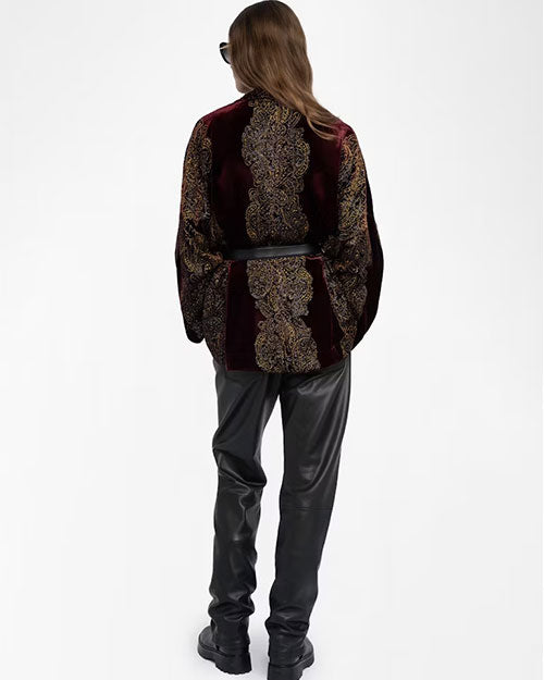 Back view of an outfit featuring a deep burgundy velvet kimono-style jacket with an intricate gold and black paisley pattern. The jacket is cinched at the waist with a black belt and paired with black leather pants and black boots. The person is also wearing dark sunglasses, and the jacket’s design showcases a vertical ornate motif running down the center of the back.