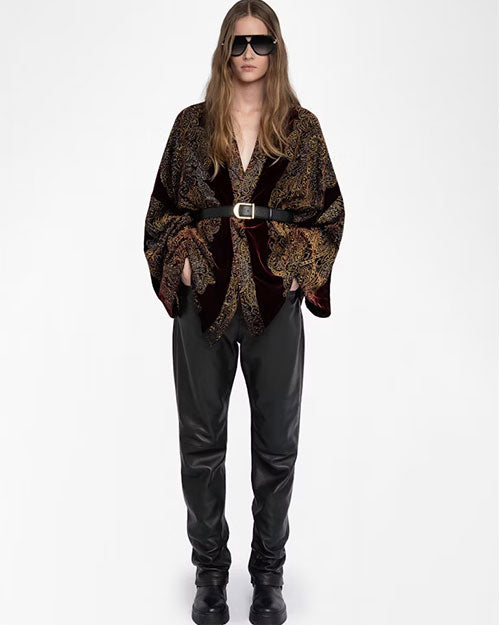 A person wearing a deep burgundy velvet kimono-style jacket with an intricate gold and black paisley pattern. The jacket has wide sleeves and is cinched at the waist with a black belt featuring a gold buckle. It is paired with black leather pants and black boots. The outfit combines rich textures and a bold, elegant design.