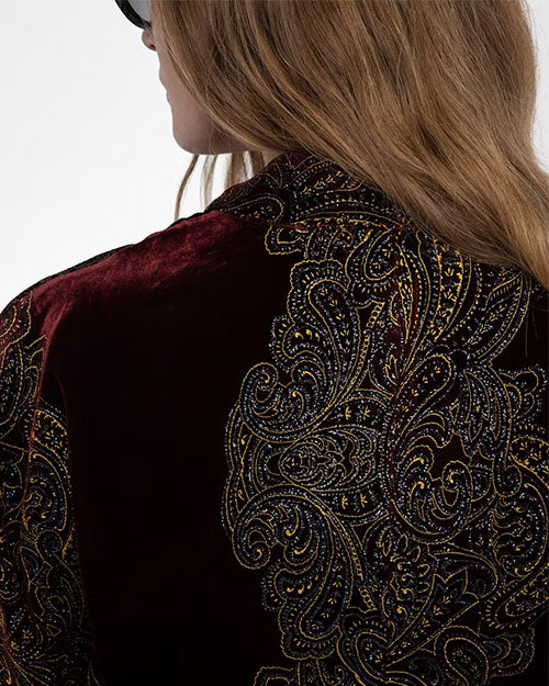 Close-up of the back of a garment made from deep burgundy velvet fabric, featuring an intricate paisley pattern embroidered in metallic gold and black thread. The design is ornate and detailed, covering a large portion of the fabric, creating a luxurious and textured appearance. The person wearing the garment has long, wavy brown hair partially visible in the frame.