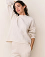 Cream-colored cable-knit sweater with long sleeves and a ribbed crew neckline, paired with matching knit bottoms. The sweater features a relaxed fit and a small decorative emblem near the hem.