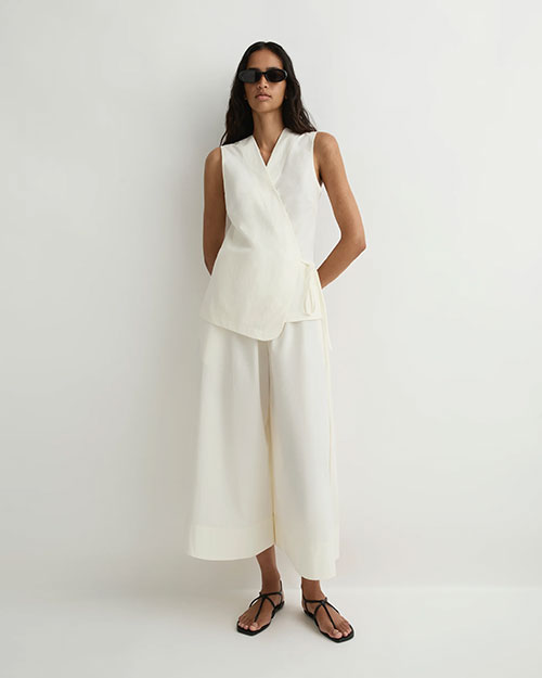 Person modeling an ivory sleeveless wrap-style top with a side tie and matching wide-leg cropped pants, paired with black flat sandals and sunglasses against a plain light background.