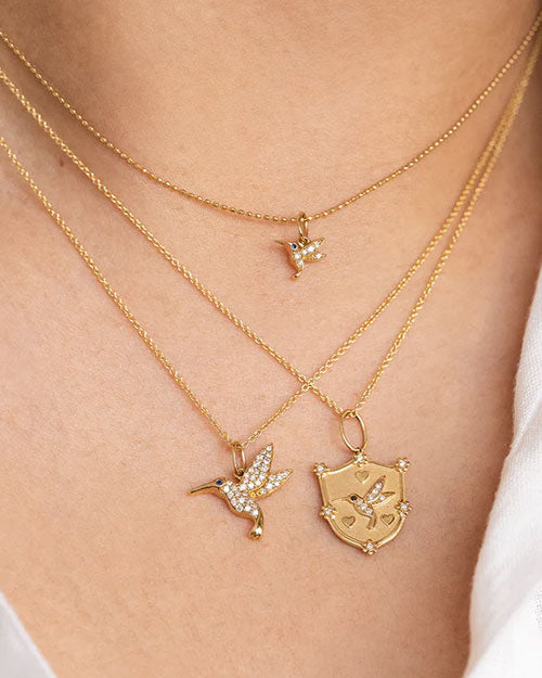 Three gold necklaces worn around a neck, each featuring a unique pendant. The top necklace has a small bird-shaped pendant with blue and white stones. The middle necklace displays a larger bird-shaped pendant adorned with white stones and a blue stone for the eye. The bottom necklace features a shield-shaped pendant with an engraved bird design and decorative elements at the corners.