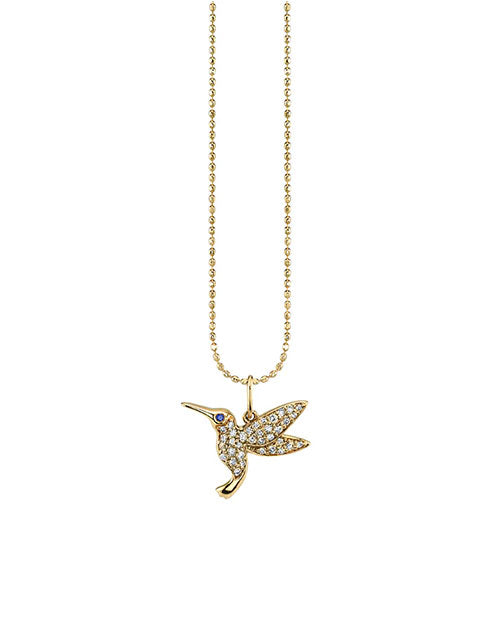 A gold necklace featuring a hummingbird-shaped pendant adorned with small sparkling stones. The body and wings are encrusted with clear stones, and the eye is represented by a small blue stone. The chain is made of uniformly spaced round links.