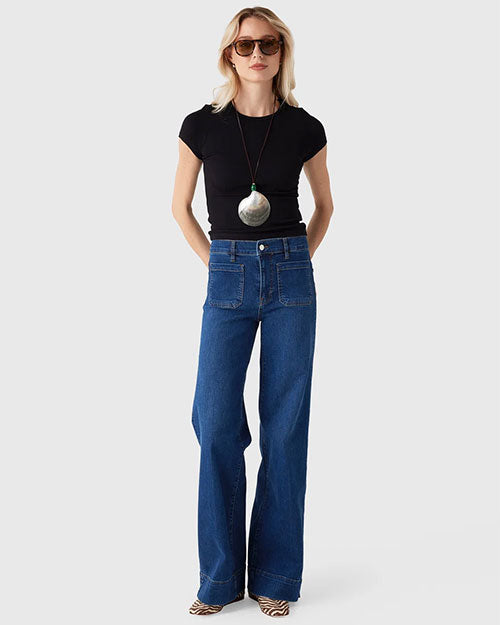 Person modeling a fitted black short-sleeve top tucked into high-waisted wide-leg blue jeans. The outfit is accessorized with oversized brown sunglasses and a long necklace featuring a large round silver pendant with a green accent. The person stands against a light gray background with hands behind the back and wears patterned shoes visible beneath the jeans.