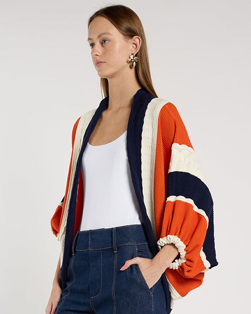 Person standing in a studio against a light neutral background, wearing a white scoop-neck tank top layered under a cropped, open-front cardigan with textured color-block stripes in rust orange, navy, and cream.