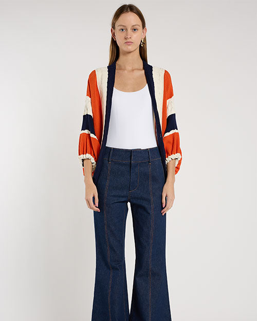 Person standing in a studio against a light neutral background, wearing a white scoop-neck tank top layered under a cropped, open-front cardigan with textured color-block stripes in rust orange, navy, and cream.