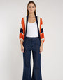 Person standing in a studio against a light neutral background, wearing a white scoop-neck tank top layered under a cropped, open-front cardigan with textured color-block stripes in rust orange, navy, and cream.