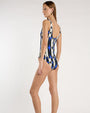 Back view of a person standing against a light neutral background, wearing a one piece swimsuit with a scoop neckline and a bold geometric pattern in blue, black, beige, and white. 