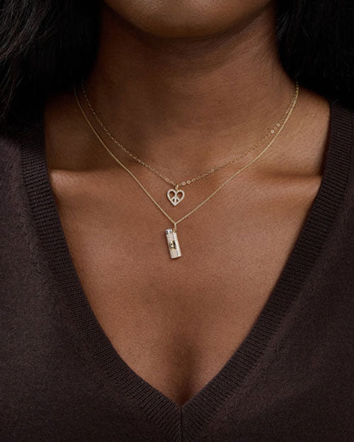 Close-up of a neck wearing two gold necklaces. One has a hollow heart-shaped pendant, and the other features a cylindrical charm with intricate detailing.