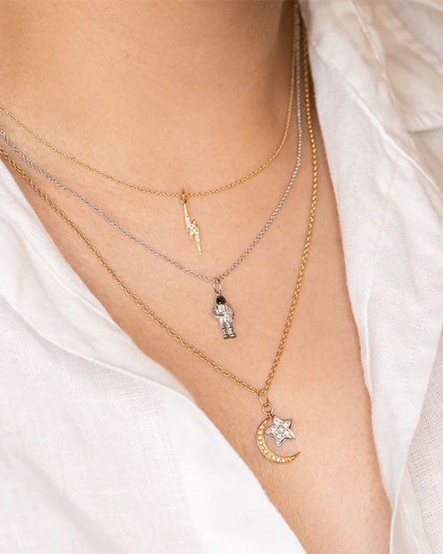 Close-up of three layered necklaces over a white shirt: a gold chain with a lightning bolt pendant, a silver chain with an astronaut pendant, and a gold chain with a crescent moon and star pendant adorned with small stones.