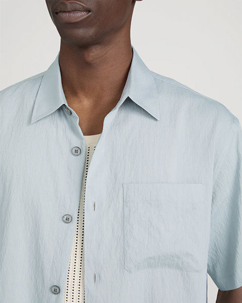 Close-up studio image against a light neutral background, showing a light blue short-sleeve button-down shirt with a pointed collar and visible front buttons. The image highlights the upper chest area, including a single chest pocket and the textured fabric. A cream ribbed tank top is visible underneath, layered beneath the open shirt.