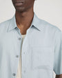Close-up studio image against a light neutral background, showing a light blue short-sleeve button-down shirt with a pointed collar and visible front buttons. The image highlights the upper chest area, including a single chest pocket and the textured fabric. A cream ribbed tank top is visible underneath, layered beneath the open shirt.