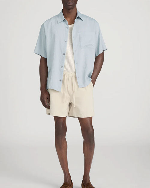 Person standing in a studio against a light neutral background, wearing a light blue short-sleeve button-down shirt layered open over a cream ribbed tank top. The outfit is paired with beige drawstring shorts and brown slip-on shoes, with one hand placed in a pocket, photographed in a full-length, front-facing pose.