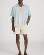 Person standing in a studio against a light neutral background, wearing a light blue short-sleeve button-down shirt layered open over a cream ribbed tank top. The outfit is paired with beige drawstring shorts and brown slip-on shoes, with one hand placed in a pocket, photographed in a full-length, front-facing pose.