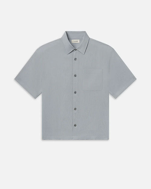 Product image of a light blue short-sleeve button-down shirt displayed flat against a light background, featuring a pointed collar, a full button front with tonal buttons, a single chest pocket, and a subtle textured fabric. The shirt has a clean, relaxed silhouette with straight hem.