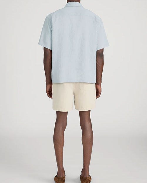 Rear-view studio image of a person standing against a light neutral background, wearing a light blue short-sleeve button-down shirt with a relaxed fit and straight hem. The outfit is paired with beige shorts that fall above the knee and brown slip-on shoes, photographed in a full-length, back-facing pose.