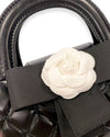 Close-up of a black quilted handbag with rounded top handles, featuring a large white flower embellishment and a black grosgrain bow.