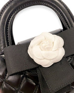 Close-up of a black quilted handbag with rounded top handles, featuring a large white flower embellishment and a black grosgrain bow.