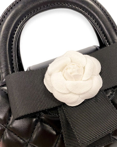 Close-up of a black quilted handbag with rounded top handles, featuring a large white flower embellishment and a black grosgrain bow.