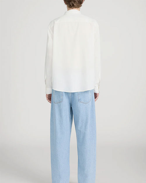 Rear-view studio image of a person standing against a light neutral background, wearing a lightweight off-white long-sleeve button-down shirt with a relaxed fit and a curved hem. The outfit is paired with light-wash wide-leg jeans, photographed in a full-length, back-facing pose.