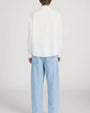 Rear-view studio image of a person standing against a light neutral background, wearing a lightweight off-white long-sleeve button-down shirt with a relaxed fit and a curved hem. The outfit is paired with light-wash wide-leg jeans, photographed in a full-length, back-facing pose.