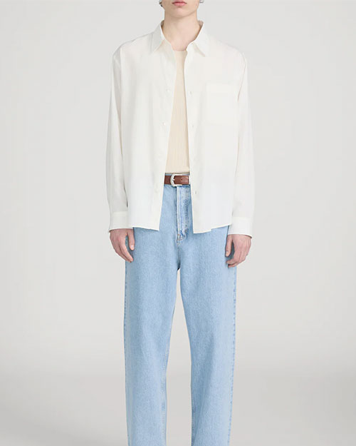 Person standing in a studio against a light neutral background, wearing a lightweight off-white long-sleeve button-down shirt left open over a beige crewneck top. The outfit is paired with light-wash straight-leg jeans and a brown leather belt, photographed in a full-length, front-facing pose.