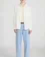 Person standing in a studio against a light neutral background, wearing a lightweight off-white long-sleeve button-down shirt left open over a beige crewneck top. The outfit is paired with light-wash straight-leg jeans and a brown leather belt, photographed in a full-length, front-facing pose.