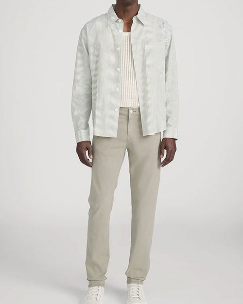 Person standing in a studio against a light neutral background, wearing a light striped button-down shirt with long sleeves left open over a cream ribbed tank top. The outfit is paired with slim-fit khaki pants and white lace-up sneakers, photographed in a full-length, front-facing pose.