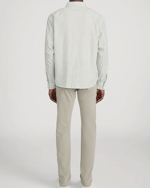 Rear-view studio image of a person standing against a light neutral background, wearing a long-sleeve button-down shirt with fine vertical stripes in light neutral tones. The shirt has a relaxed fit and a curved hem. The outfit is paired with slim-fit khaki pants and white lace-up sneakers, photographed in a full-length, back-facing pose.