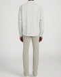 Rear-view studio image of a person standing against a light neutral background, wearing a long-sleeve button-down shirt with fine vertical stripes in light neutral tones. The shirt has a relaxed fit and a curved hem. The outfit is paired with slim-fit khaki pants and white lace-up sneakers, photographed in a full-length, back-facing pose.
