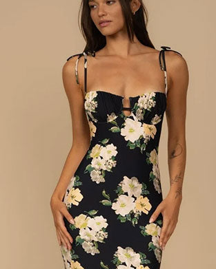 Person wearing a fitted, sleeveless black dress with thin tie straps and a large floral print featuring white and light yellow flowers with green leaves.