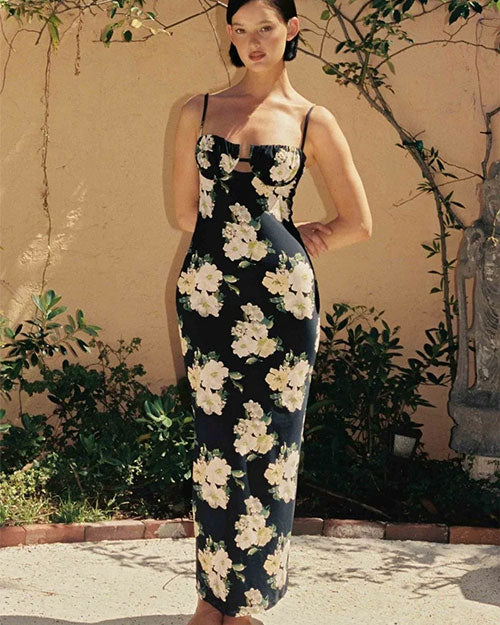 Person wearing a long, fitted black dress with thin spaghetti straps and a large white floral print, standing outdoors against a beige wall with greenery in the background.