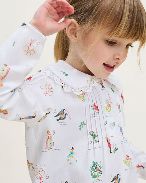 Young child wearing a white shirt with a lace collar and pleated front, decorated with colorful illustrations and numbers including birds, people, and objects. The child is looking downward with one hand raised near their head.