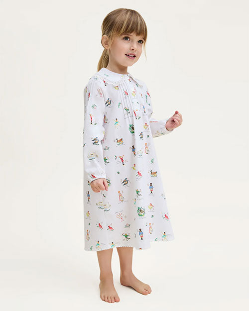  Young child standing barefoot against a plain background, wearing a long-sleeved white dress adorned with colorful illustrations of small figures and animals. The child’s hair is tied back, highlighting the playful and whimsical design of the dress.