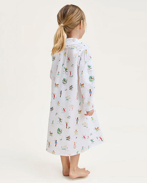 Child standing barefoot with their back to the camera, wearing a long-sleeved white nightgown decorated with colorful illustrations. The child’s hair is tied back in a ponytail, and the background is plain and light-colored.